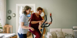 Comment bien choisir votre vélo d’appartement pour séniors ? Mature female physical therapist assisting elderly woman on exercise bike at nursing home