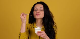 Consommer du yaourt le soir : est-ce conseillé ou non ? Beautiful young woman enjoying eating a freshly yogurt while standing against yellow background. Healthy breakfast concept. Real time