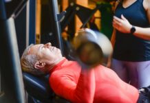 Sport santé : 5 conseils pour optimiser votre bien-être Homme senior effectuant un exercice de musculation avec haltères sur un banc de musculation, encadré par une coach sportive professionnelle pour une pratique du sport santé sécurisée.