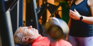 Sport santé : 5 conseils pour optimiser votre bien-être Homme senior effectuant un exercice de musculation avec haltères sur un banc de musculation, encadré par une coach sportive professionnelle pour une pratique du sport santé sécurisée.
